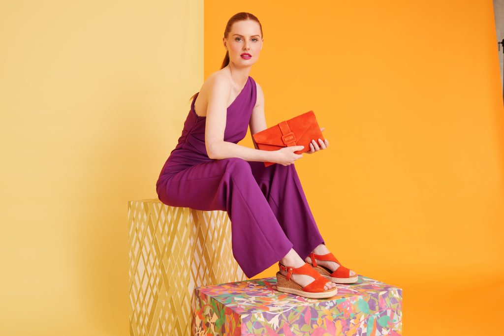 Woman wears a purple jumpsuit with contrasting orange sandals and clutch bag